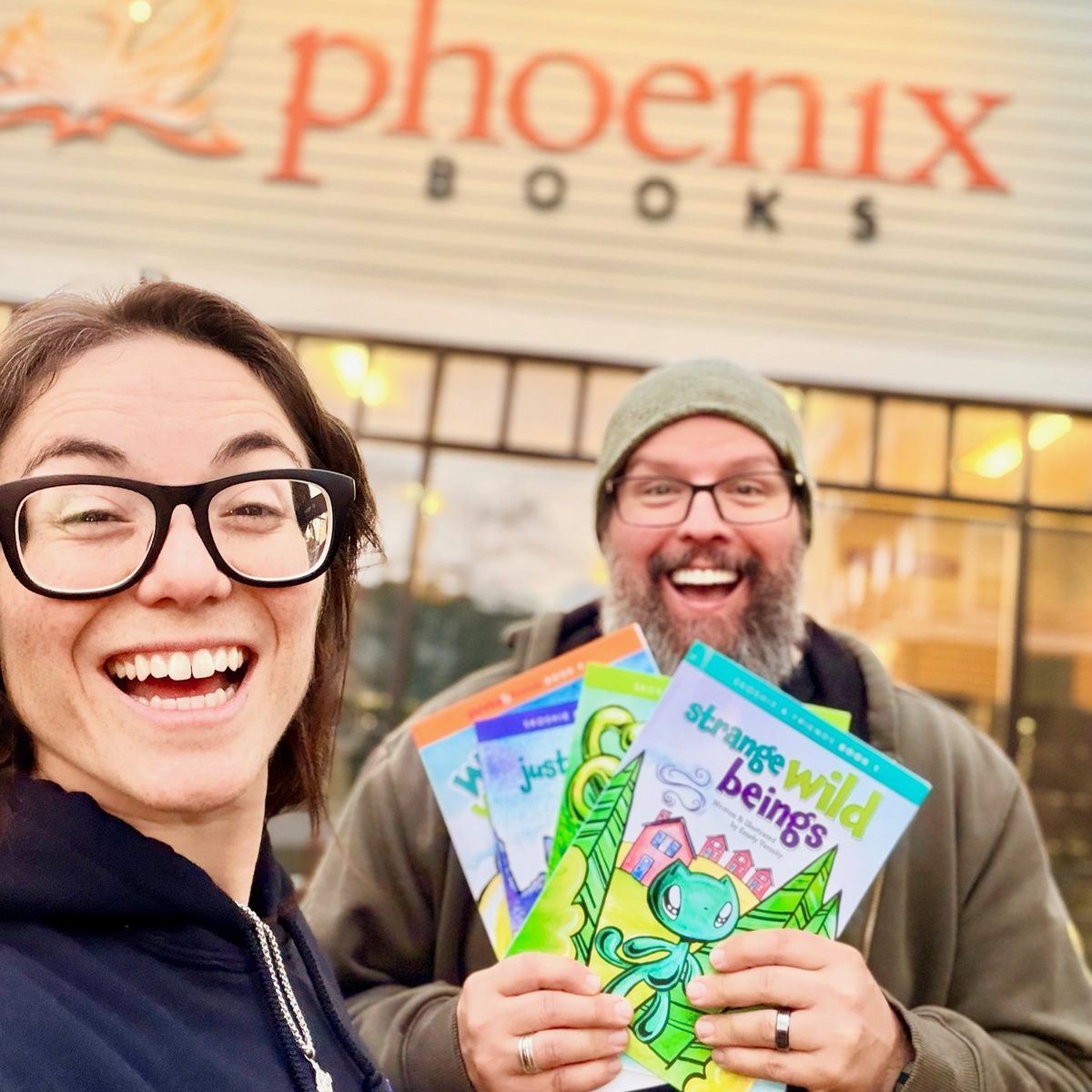 Two smiling people holding Skoshie & Friends picture books in front of the Phoenix Books store sign in Essex Junction, Vermont